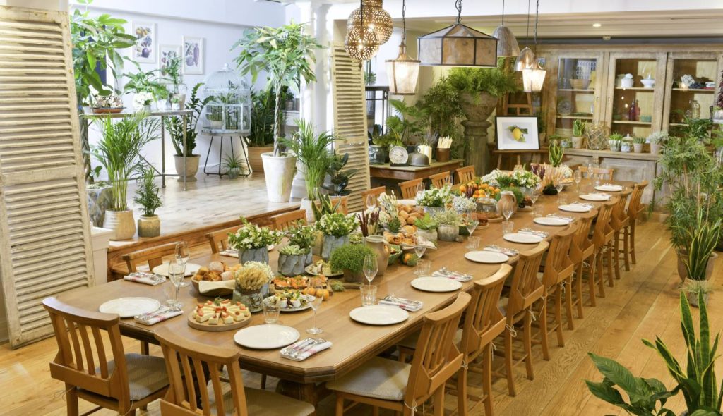 Daytime view of Conservatory with long table laid with plates, cutlery, Prosecco glasses, fruit platters, and small tubs of flowers.