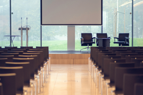 Image from the back of an empty-theatre layout conference room with a stage, lectern, and chairs for a speaker panel. There are floor-to-ceiling windows behind the stage, filling the room with natural light.