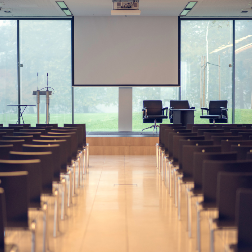 Image from the back of an empty-theatre layout conference room with a stage, lectern, and chairs for a speaker panel. There are floor-to-ceiling windows behind the stage, filling the room with natural light.