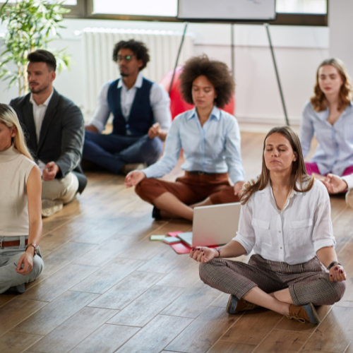 A group of people in business wear sat on a wood floor, in a basic meditation pose.