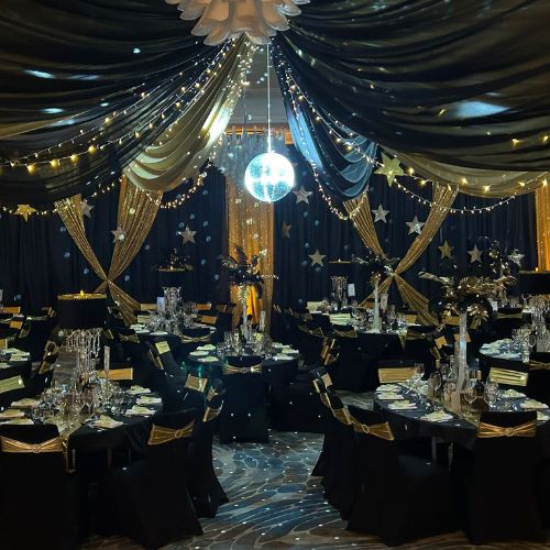 A room decorated for a corporate awards event - black table clothes and chair backs are draped in shiny gold material, lights adorn the room, and table are laid with fresh, gleaming cutlery and glassware