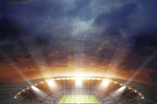 View from above an open-top sport stadium with a grass pitch and stadium lights switched on and beaming - the stadium is empty.