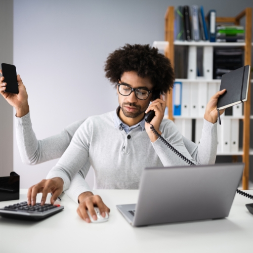 Image to represent busy PAs for whom free venue finding services for executive assistants can help - Man sat at desk multi-tasking, with extra arms using a calculator, mobile phone, keyboard, desk phone, and notebook